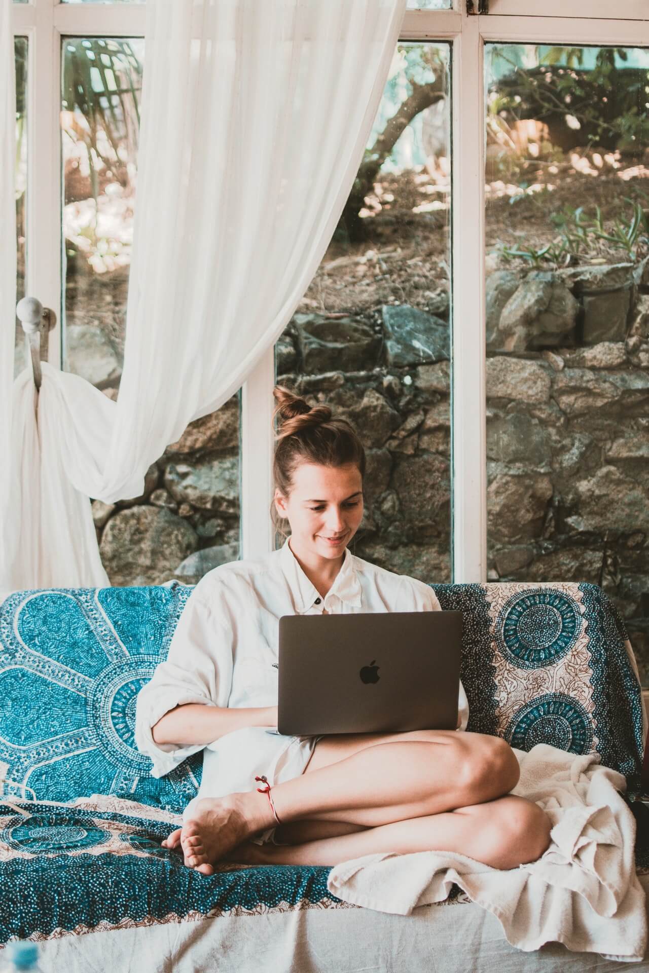 A woman sits on a blue-patterned couch, working on a laptop in a bright, cozy room with large windows and white curtains, surrounded by nature. She appears focused and comfortable, embodying a relaxed work-from-home environment, ideal for using a social media scheduler. | plannthat.com