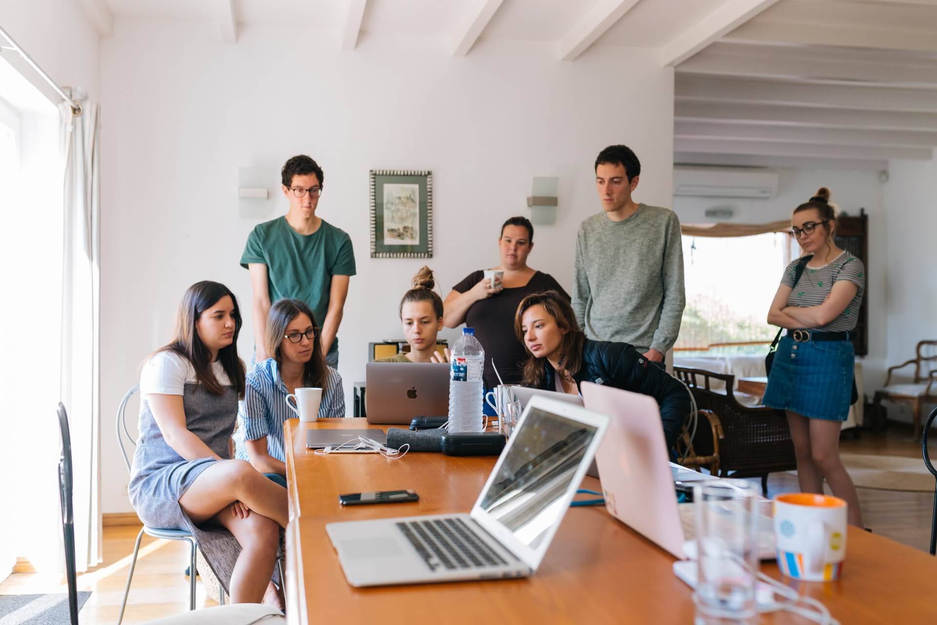 A group of seven people gathered around a table with laptops, engaged in a discussion in a bright room with wooden flooring and white walls, reviewing a social media planner | plannthat.com