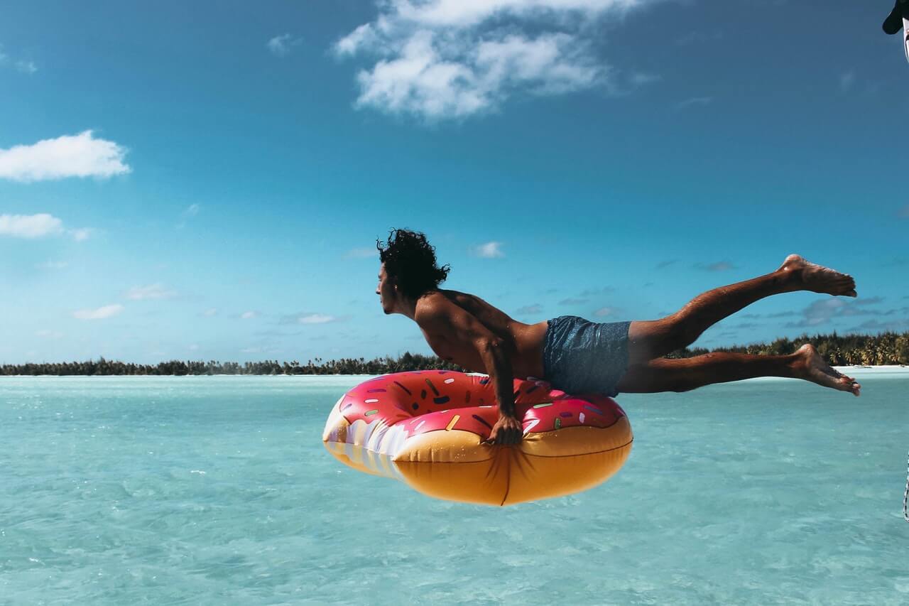 A person mid-air, diving into clear blue water while holding a colorful inflatable donut float, with a serene ocean and distant shoreline under a bright, partly cloudy sky, showcasing a perfect moment for a social media planner | plannthat.com