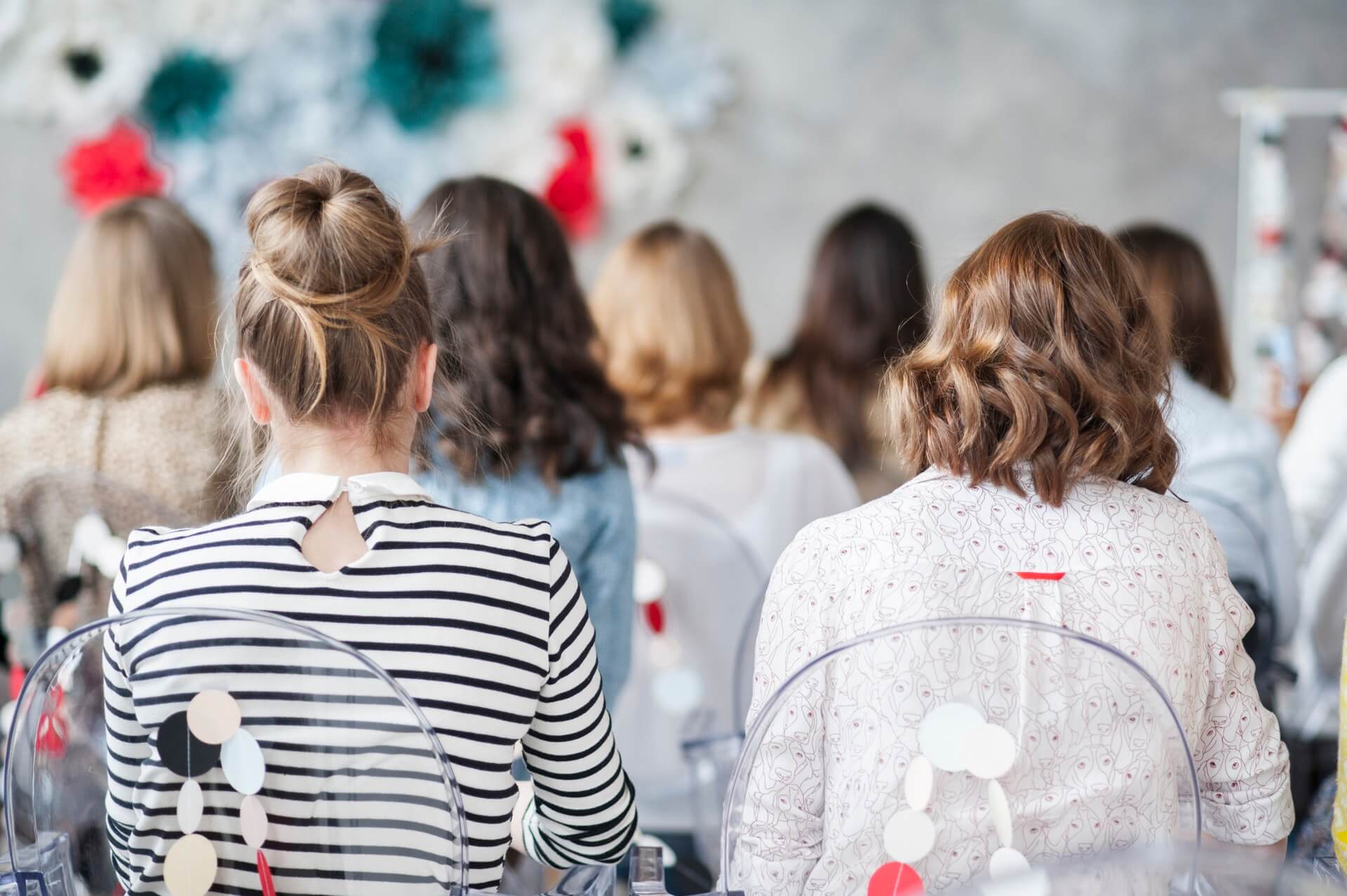 A group of women sitting indoors, viewed from behind, focused on an unseen subject, with decorated background and clear chairs, discussing strategies | Instagram scheduler | plannthat.com