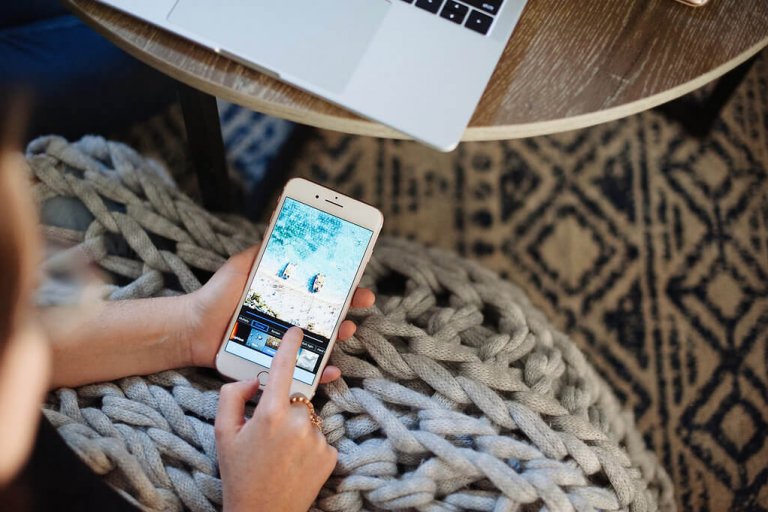 Person editing a photo on a smartphone while sitting on a chunky knit blanket, with a laptop on a nearby wooden table, using an Instagram scheduler | plannthat.com