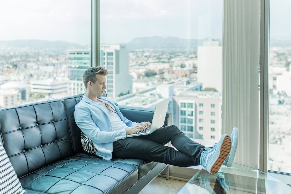 A man in a light blue blazer works on a laptop while sitting on a black leather couch in a modern high-rise apartment with large windows offering a panoramic city view, using a social media scheduler | plannthat.com