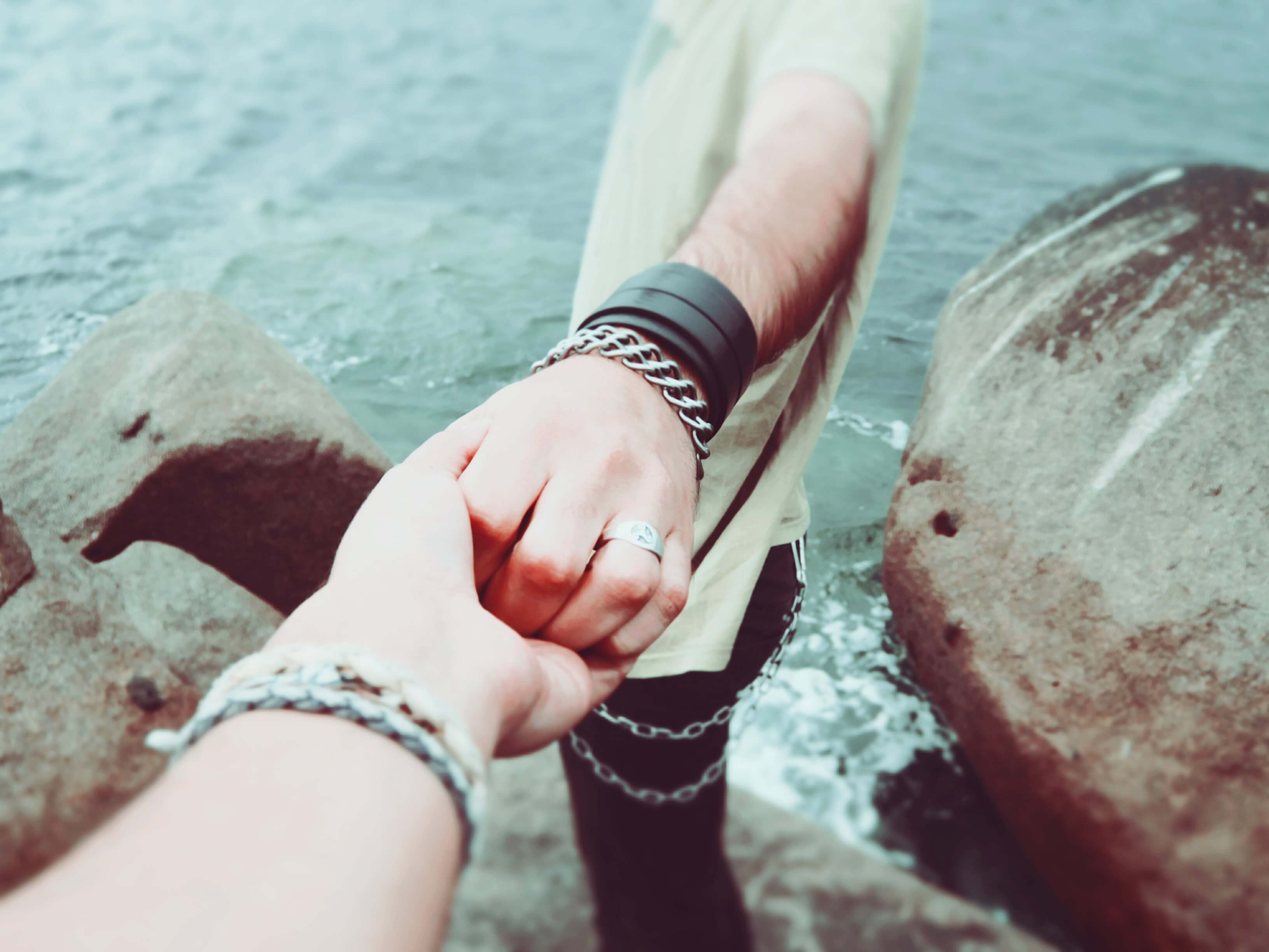 Two people holding hands near a rocky seashore, with one leading the other, wearing bracelets and rings symbolizing connection and companionship, perfect for showcasing moments on an Instagram scheduler | plannthat.com