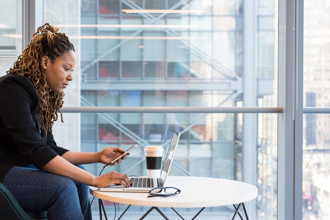 A woman with braided hair sits at a round table, using a laptop and holding a smartphone, with a coffee cup and glasses on the table in a modern office with large windows and an urban view, utilizing a social media scheduler | plannthat.com
