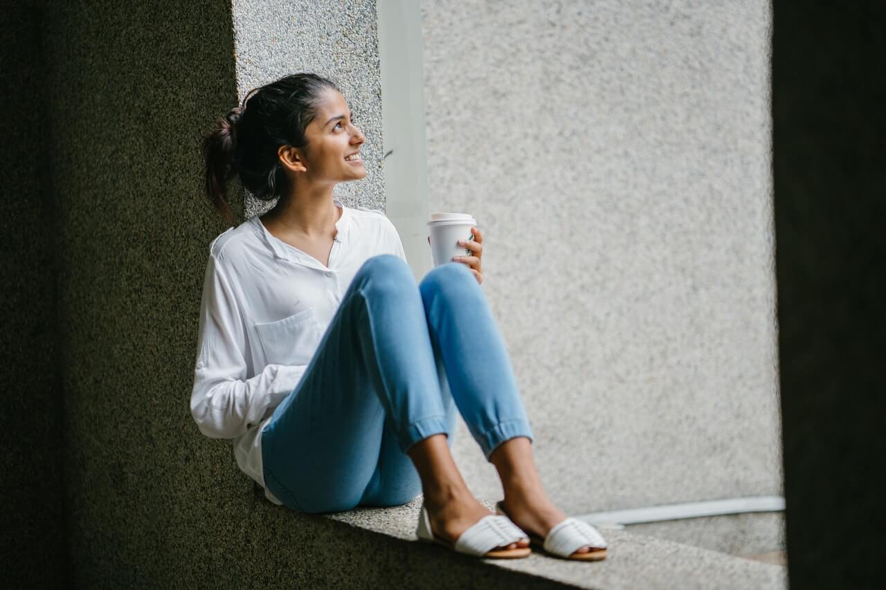 A smiling woman in a white shirt and blue jeans sits on a ledge, holding a coffee cup and looking up, representing a relaxed moment for a social media planner | plannthat.com