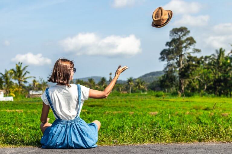 A woman sitting on the roadside in a green field, tossing a hat into the air, under a clear blue sky, enjoying her time with the help of a social media scheduler | plannthat.com
