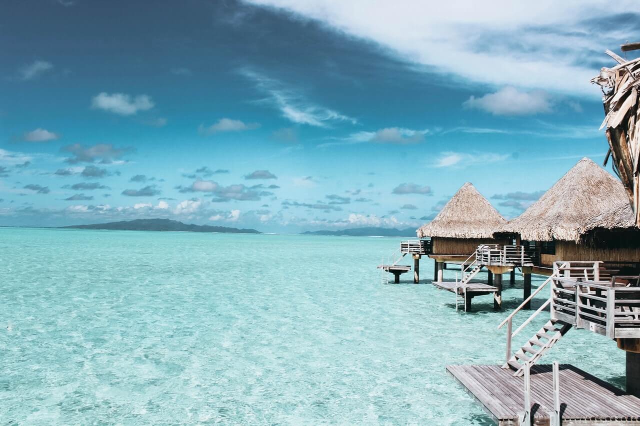 Overwater bungalows with thatched roofs above clear turquoise water under a bright blue sky with scattered clouds, with a distant island on the horizon, creating a serene and tropical vacation setting, perfect for your Instagram scheduler | plannthat.com