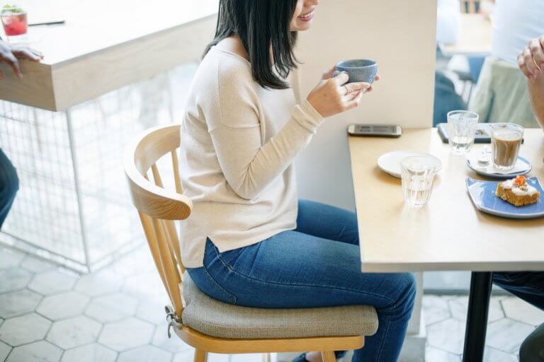Person in casual attire sitting at a café table, holding a cup, with coffee, glasses of water, a dessert plate, and a smartphone on the table; another person is partially visible in the background, illustrating a moment of planning with a social media scheduler | plannthat.com