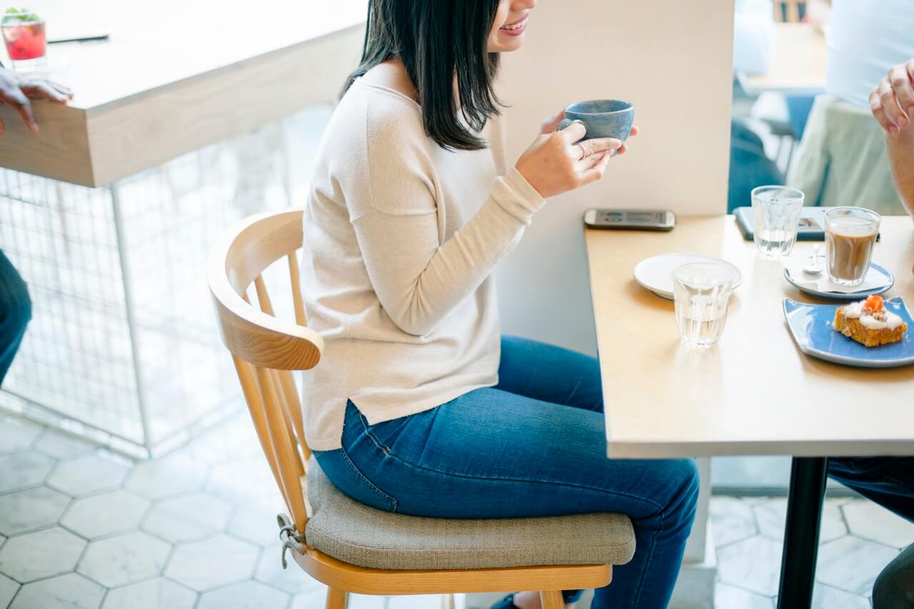 Person in casual attire sitting at a café table, holding a cup, with coffee, glasses of water, a dessert plate, and a smartphone on the table; another person is partially visible in the background, illustrating a moment of planning with a social media scheduler | plannthat.com