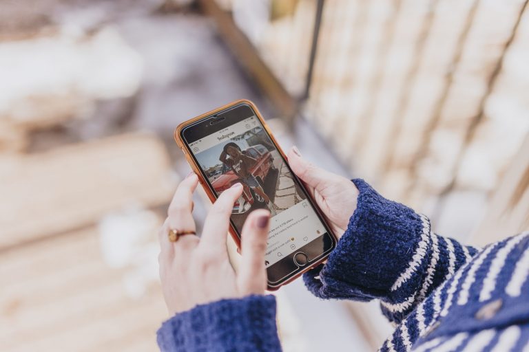 A person wearing a blue and white sweater holding a smartphone, viewing a social media post featuring a woman standing by a red car, with a blurred background of a wooden deck and some snow, using a social media scheduler | plannthat.com