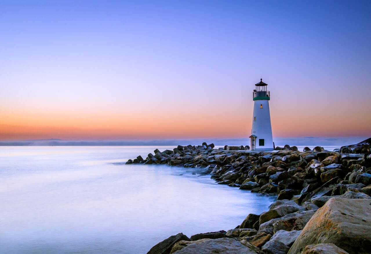 A serene seaside scene at sunset, featuring a white lighthouse on a rocky shore with a calm ocean under a gradient sky transitioning from orange to blue, perfect for a social media planner | plannthat.com