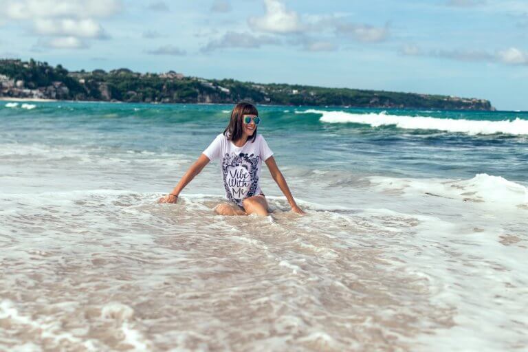 Woman in sunglasses and printed shirt sitting in shallow ocean water, enjoying the waves on a sunny day, planning her next post with a social media scheduler | plannthat.com