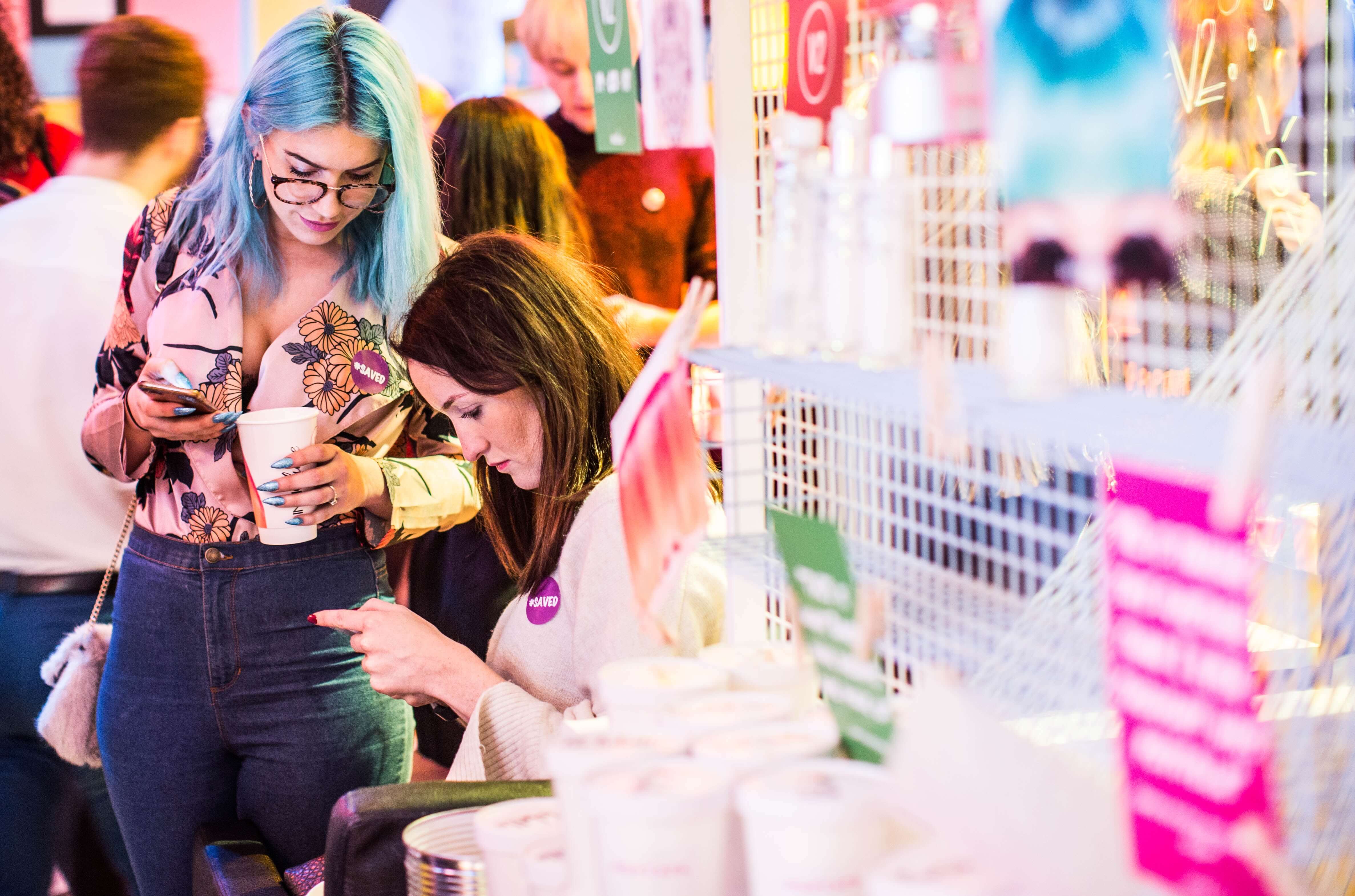 Two women at a vibrant market stall, one with blue hair holding a cup and the other pointing at something, with various products displayed and people in the background, showcasing a lively scene perfect for a social media planner | plannthat.com
