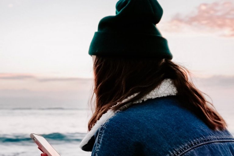 Person wearing a green beanie and denim jacket, holding a smartphone while sitting on a beach at sunset, looking at the ocean, using a social media planner | plannthat.com