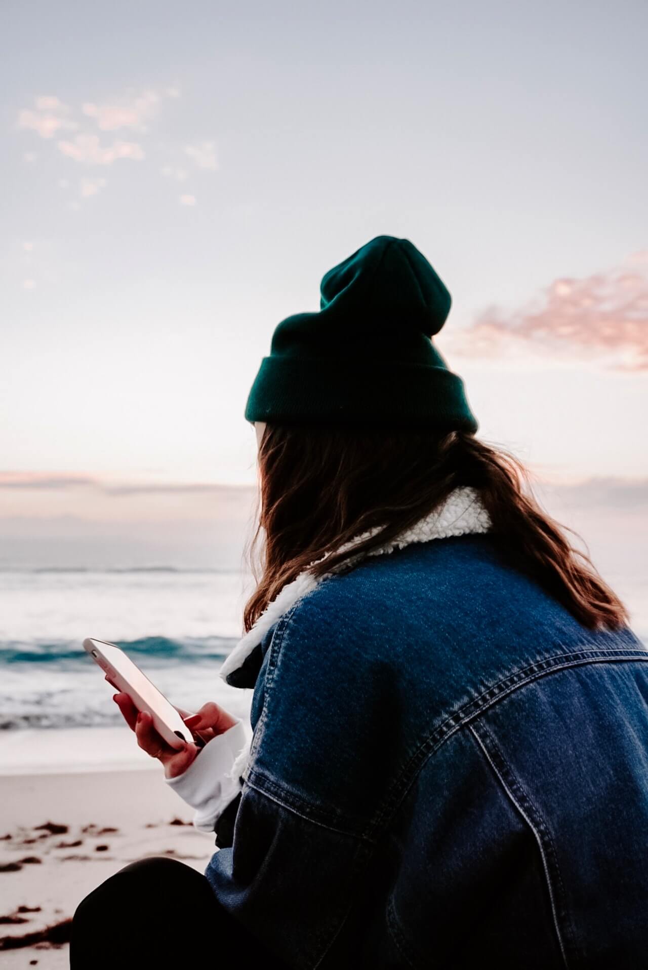 Person wearing a green beanie and denim jacket, holding a smartphone while sitting on a beach at sunset, looking at the ocean, using a social media planner | plannthat.com