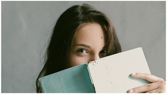 A woman with long brown hair partially hides her face behind an open book with a blue cover, revealing only her eyes, against a plain gray wall, illustrating the focus and planning needed for a social media scheduler | plannthat.com