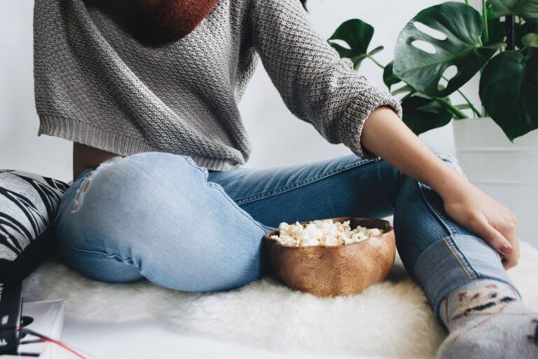 Person in cozy sweater and jeans sits cross-legged on a fluffy rug, eating popcorn from a wooden bowl, with green plants in the background, enjoying a relaxing moment while using a social media scheduler | plannthat.com