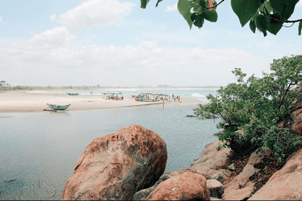 A serene beach scene with boats on the shore, people in the distance, large rocks in the foreground, and lush greenery under a partly cloudy sky, perfect for a social media scheduler | plannthat.com
