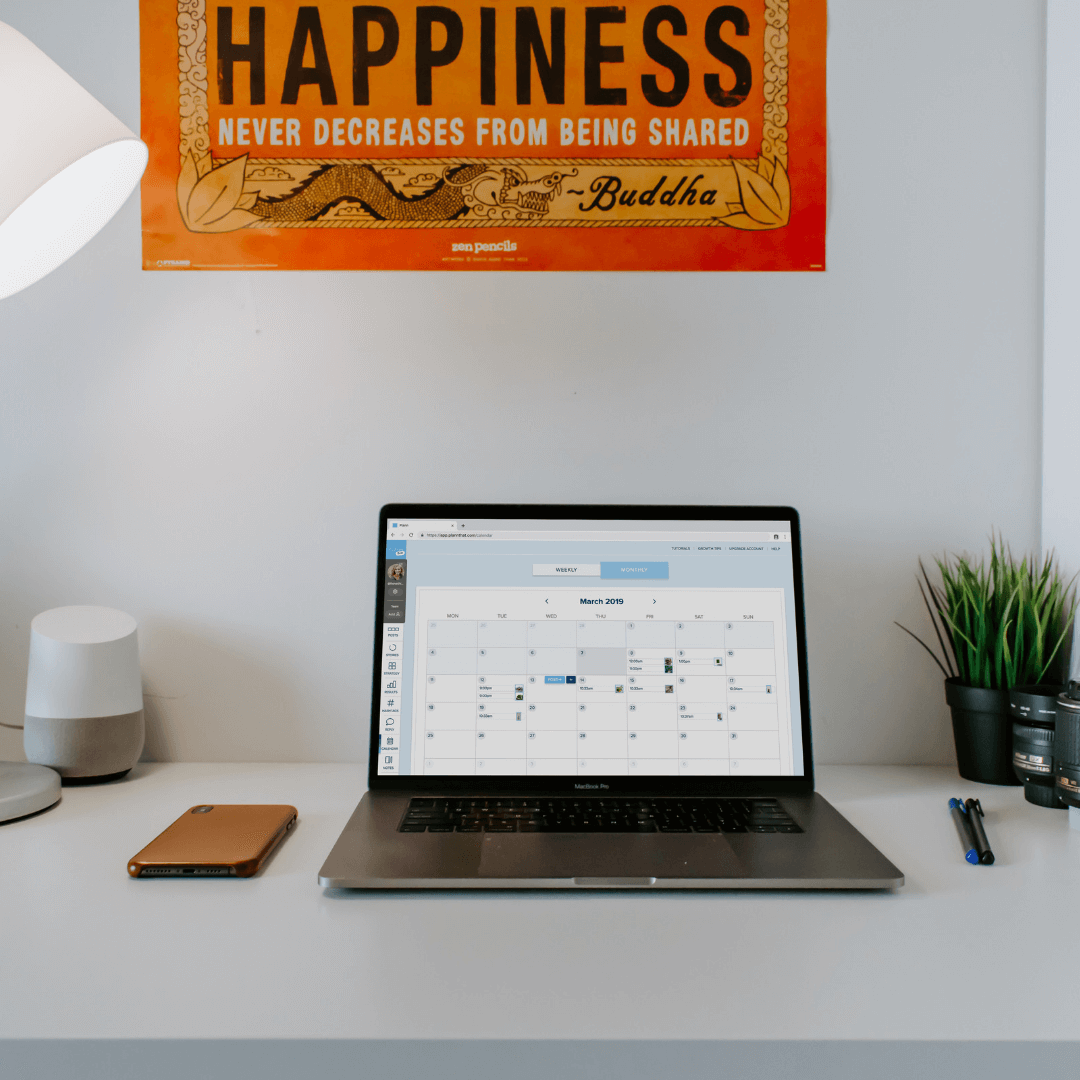 A laptop displaying a calendar sits on a desk with a phone, lamp, plant, and pens. Above the desk is a poster with the quote, "Happiness never decreases from being shared" attributed to Buddha, ideal for a social media scheduler setup | plannthat.com