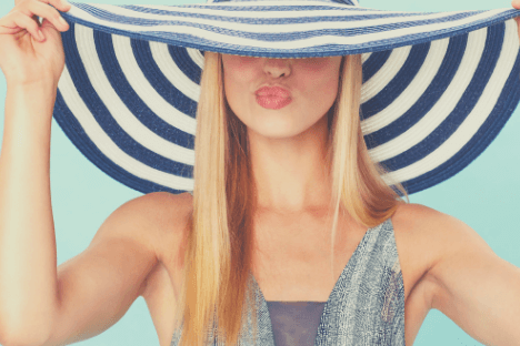 A woman with long blonde hair playfully holds a wide-brimmed, striped hat over her eyes, making a kissing face against a light blue background, showcasing a fun moment perfect for your Instagram scheduler | plannthat.com