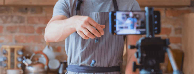 A person wearing an apron is being filmed in a kitchen with a smartphone on a tripod, likely creating a cooking video, showcasing the use of a social media scheduler | plannthat.com