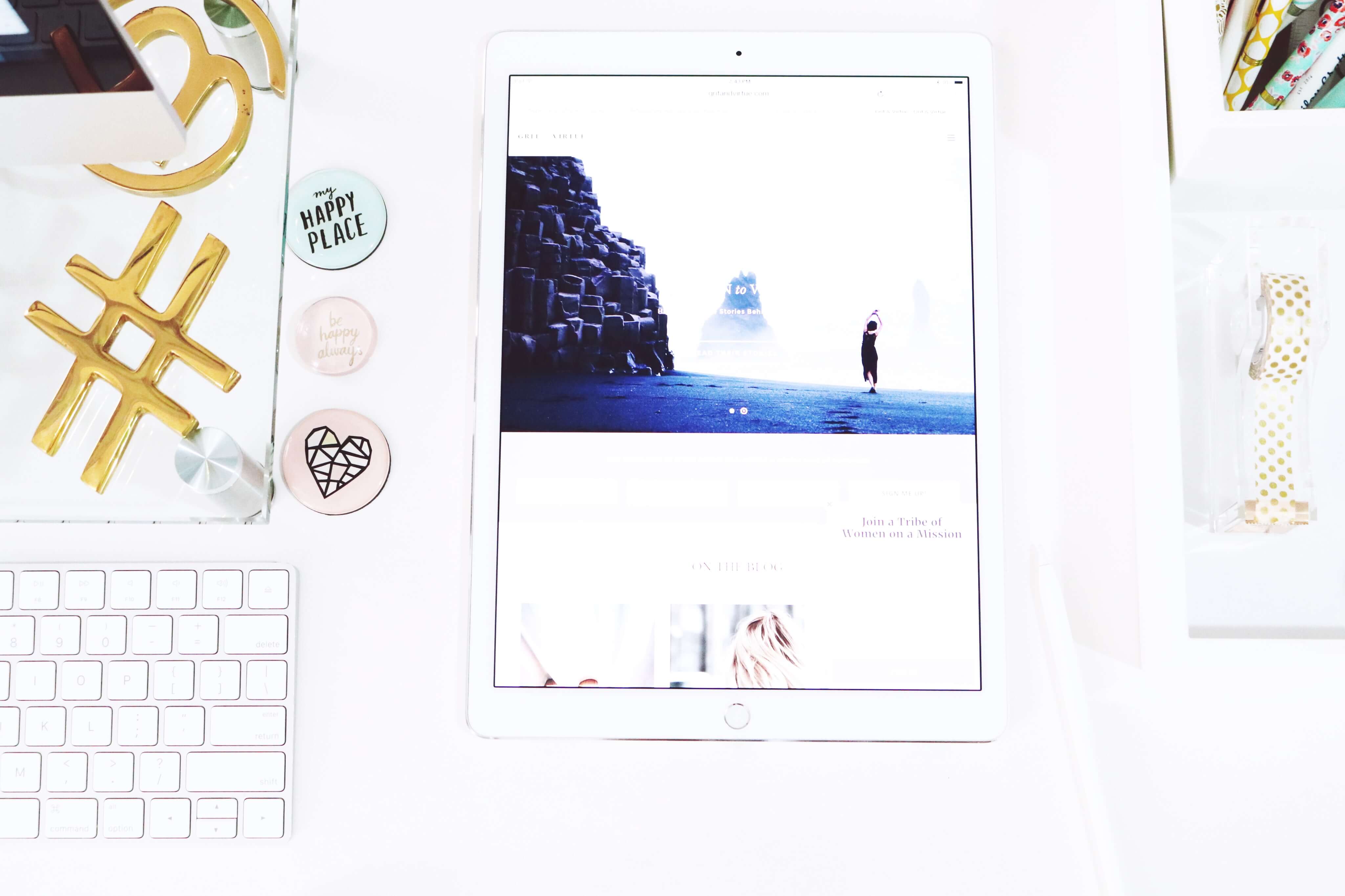 A white desk with an iPad displaying a scenic image, surrounded by a keyboard, gold hashtag decor, buttons with positive messages, and stationery items, creating a clean and organized workspace, ideal for a social media planner | plannthat.com