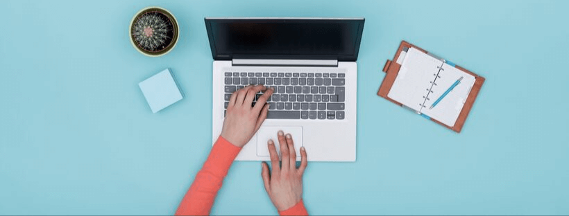 A person types on a laptop with a blue background, accompanied by a small potted cactus, a sticky note, and a clipboard with a notepad and pen, creating an organized and minimalistic scene, ideal for a social media scheduler | plannthat.com