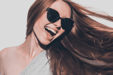 A woman with long, flowing hair and wearing sunglasses is smiling widely in a one-shoulder top, enjoying herself with her hair blowing to the side against a light background | Social media scheduler | plannthat.com