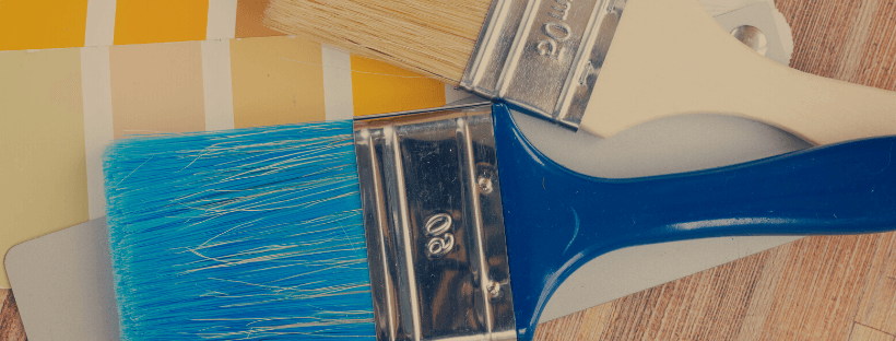 Close-up of two paintbrushes, one with blue bristles and the other with beige bristles, on a wooden surface next to a color swatch with yellow and gray shades, ideal for a social media planner | plannthat.com