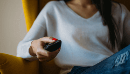 A person in a white shirt and blue jeans sitting on a yellow chair, holding a TV remote control with focus on the hand | Social media scheduler | plannthat.com