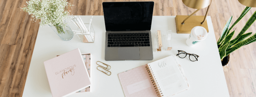 A tidy workspace featuring a laptop, open planner, glasses, coffee cup, gold paperclips, a pink book titled "Glamorous Planning," a vase with white flowers, and a potted plant on a white desk with a wooden floor background, ideal for using a social media planner | plannthat.com