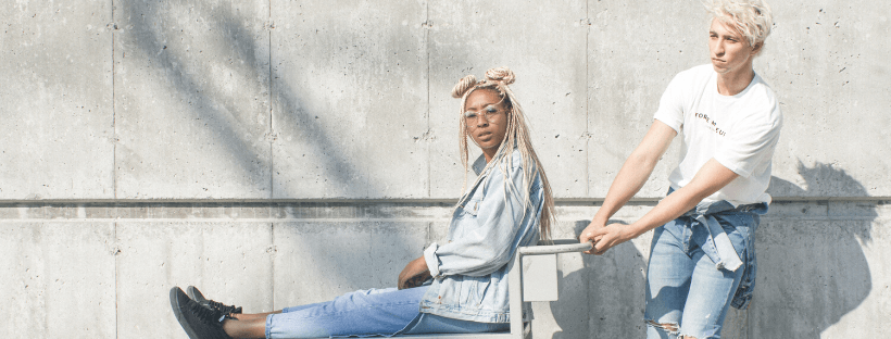 Two people in casual denim outfits, one sitting on a bench and the other leaning against it, with a concrete wall background, illustrating a relaxed moment | Social media scheduler | plannthat.com