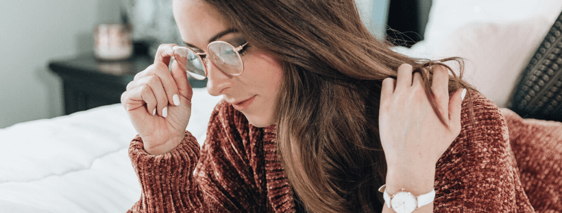 Woman with glasses and long hair wearing a red sweater, sitting on a bed, looking down thoughtfully with a wristwatch visible on her hand, blurred background includes a bed and nightstand, perfect moment for using a social media scheduler | plannthat.com