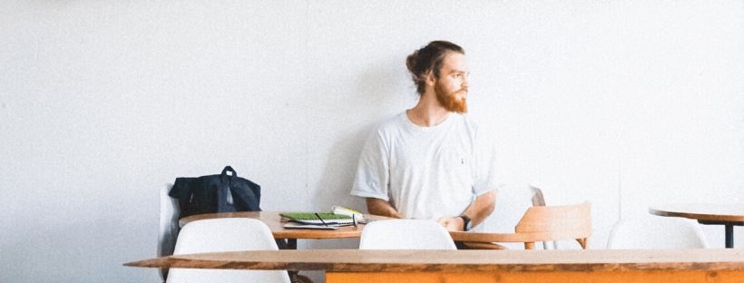 A man with a beard and a bun sits alone at a table in a minimalist room, wearing a white shirt and looking to the side. A backpack and notebooks are on the table, suggesting he might be planning his next steps with a social media scheduler | plannthat.com