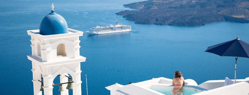 A woman relaxes in a rooftop pool overlooking the blue sea and a cruise ship in Santorini, Greece, with a white bell tower and blue dome in the background, shaded by an umbrella, perfect for your Instagram scheduler | plannthat.com
