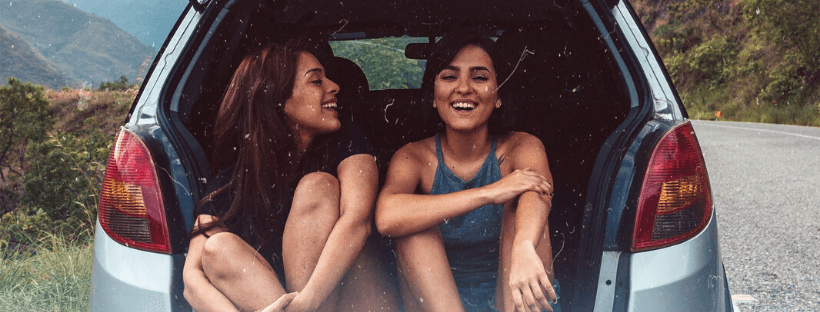 Two women sitting in a car's open trunk, laughing and enjoying a fun road trip with a scenic, mountainous landscape in the background | Social media scheduler | plannthat.com