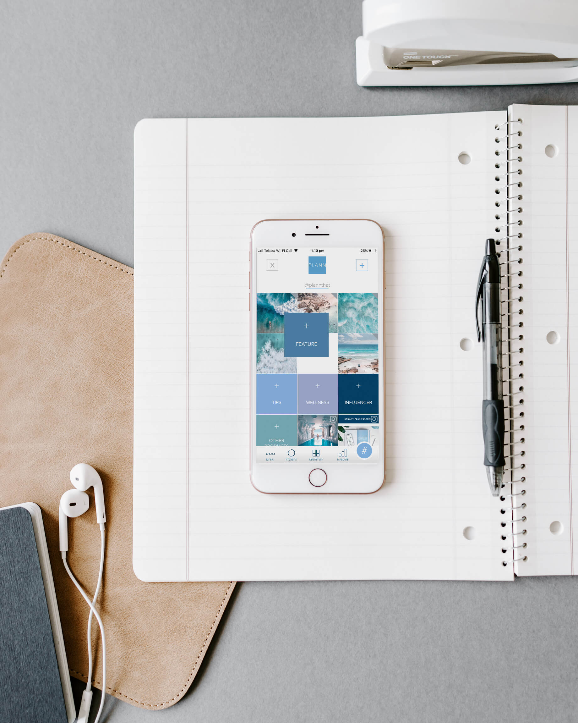 An iPhone displaying a social media scheduler app rests on an open notebook with a pen. Nearby are a leather case, earphones, and another notebook, all on a gray surface, creating an organized workspace. | plannthat.com