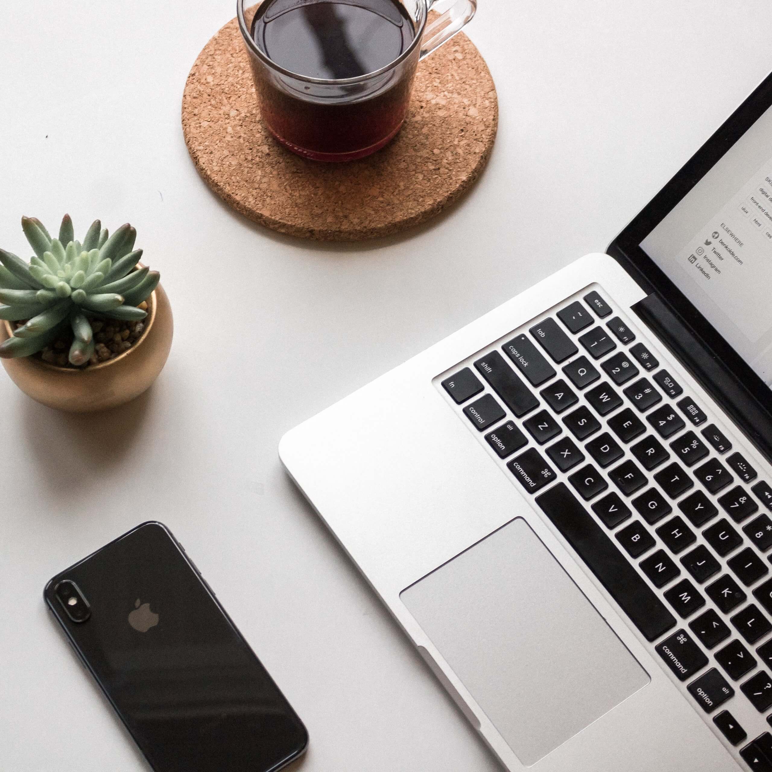 A workspace with a laptop, a smartphone, a small succulent plant, and a cup of coffee on a cork coaster, all placed on a white desk, ideal for using a social media scheduler | plannthat.com