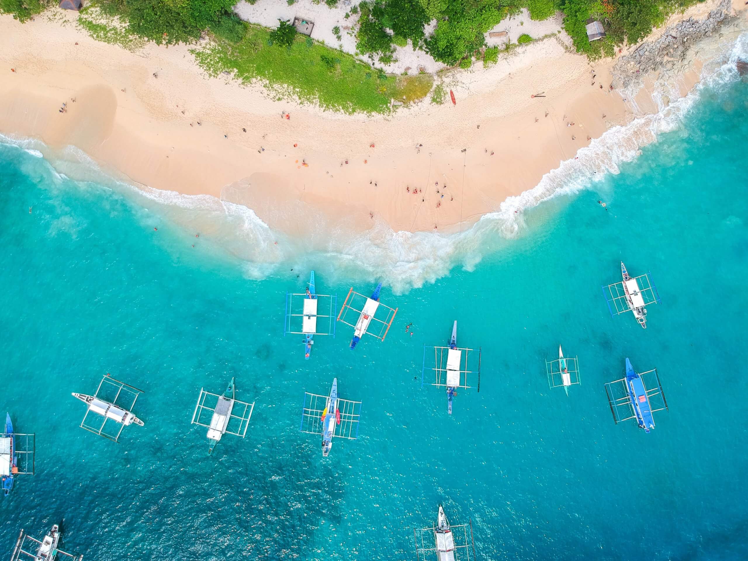 Aerial view of a sandy beach with people relaxing and swimming in clear turquoise waters, alongside multiple boats anchored near the shore, bordered by lush greenery, perfect for sharing on Instagram scheduler | plannthat.com