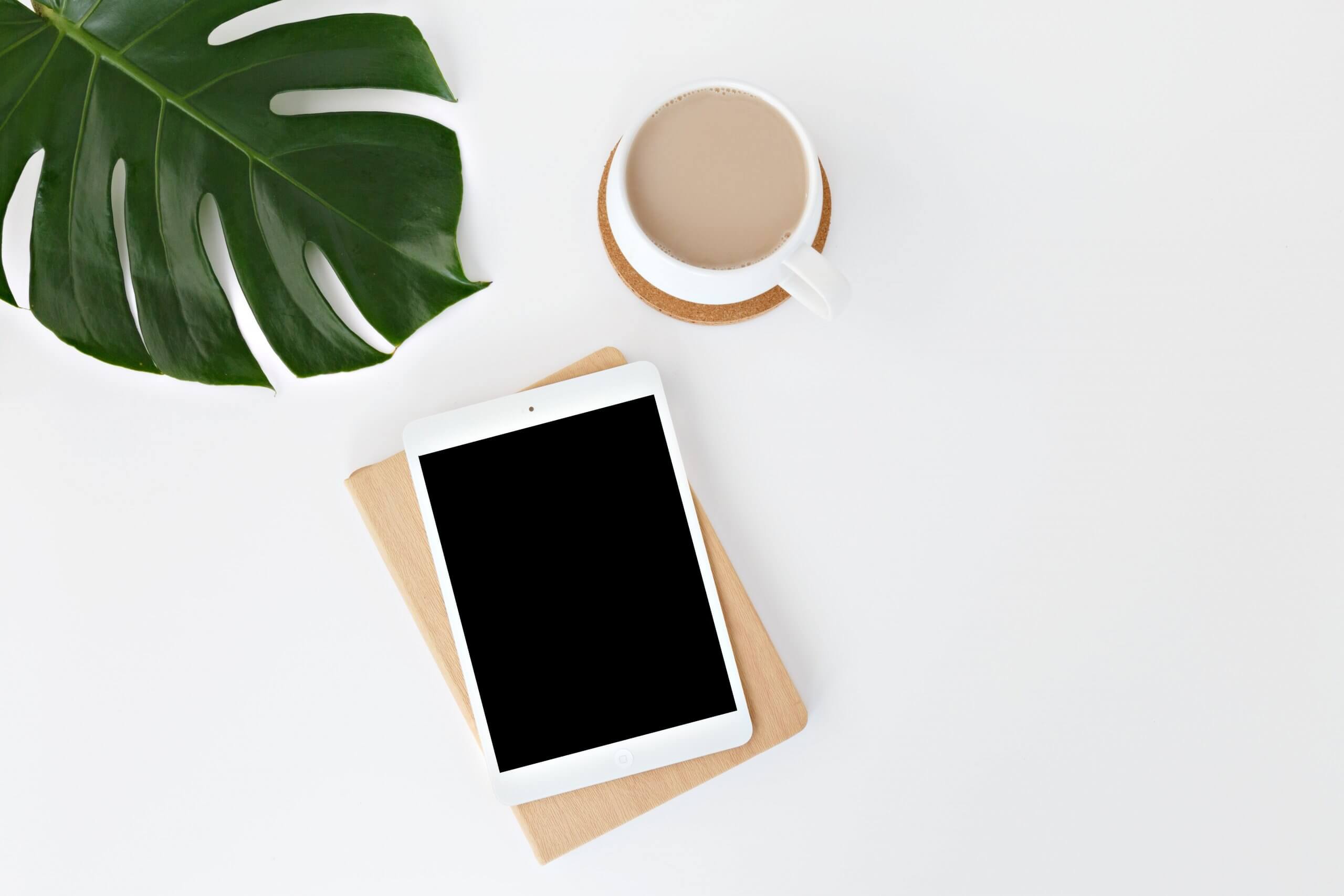 A white iPad with a black screen on a wooden surface beside a cup of coffee and a large green monstera leaf, creating a minimalistic and clean scene that evokes calm and productivity, ideal for using a social media scheduler | plannthat.com