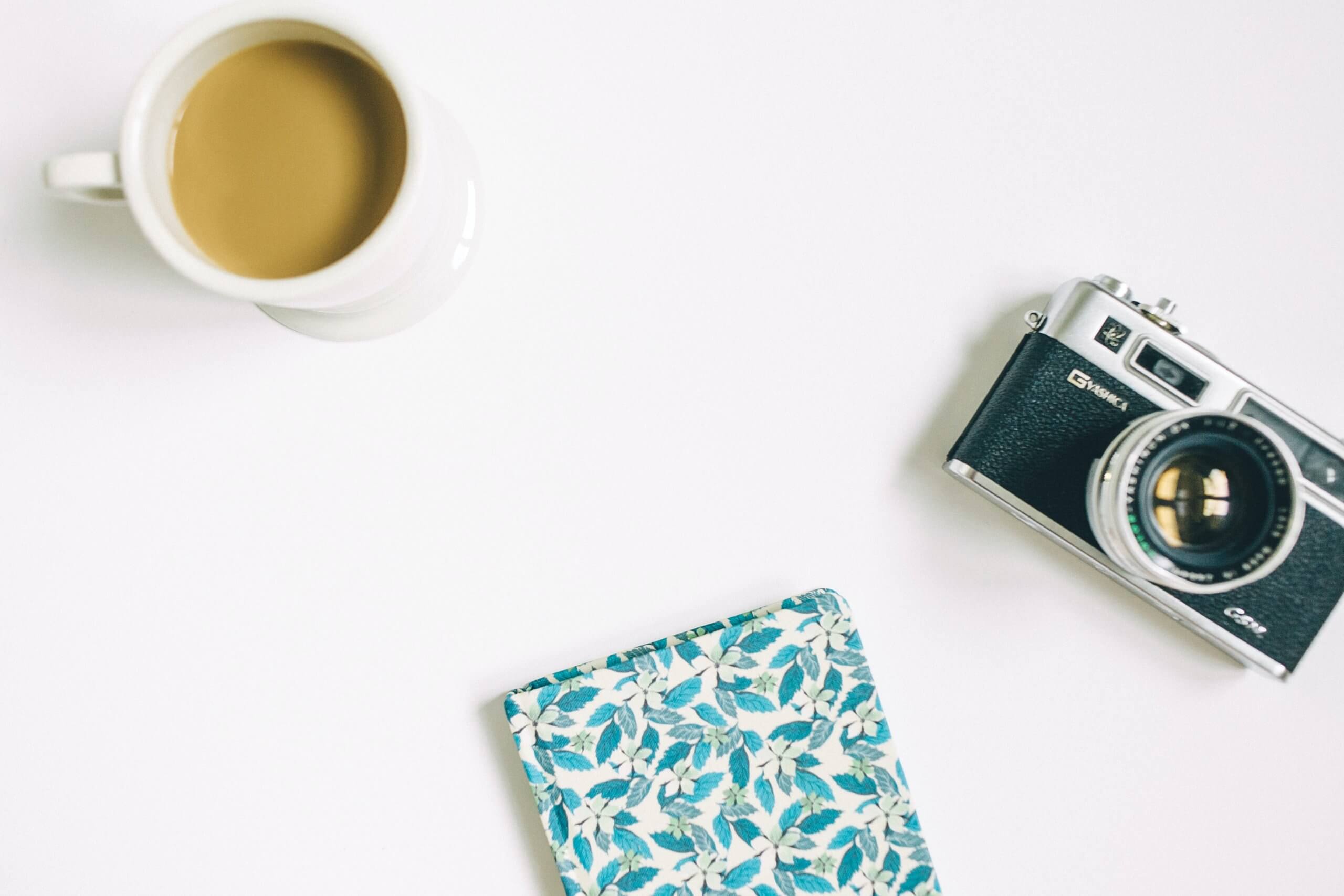 A white surface with a cup of coffee, a vintage camera, and a floral-patterned notebook, perfect for planning your social media scheduler | plannthat.com