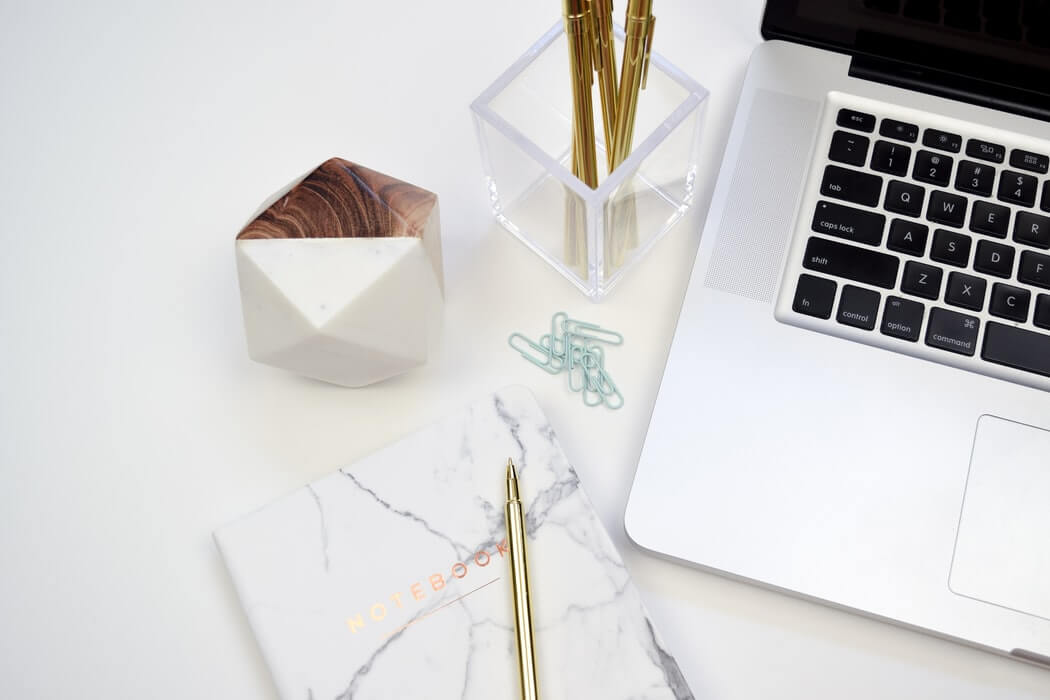A minimalist workspace featuring a laptop, marble-pattern notebook, gold pen, geometric decor, paperclips, and a clear container holding gold pens, ideal for a social media scheduler | plannthat.com