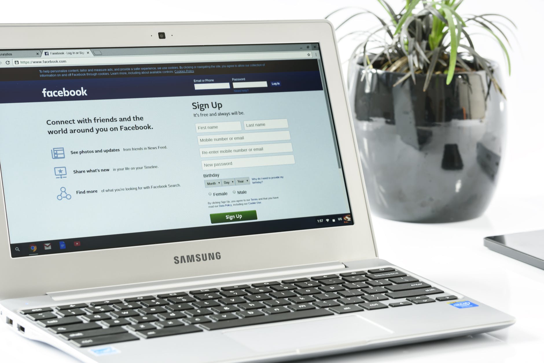 A Samsung laptop displaying the Facebook login and sign-up page is placed on a white desk, with a potted plant and part of another device visible in the background, creating a professional office setting, ideal for a social media scheduler | plannthat.com