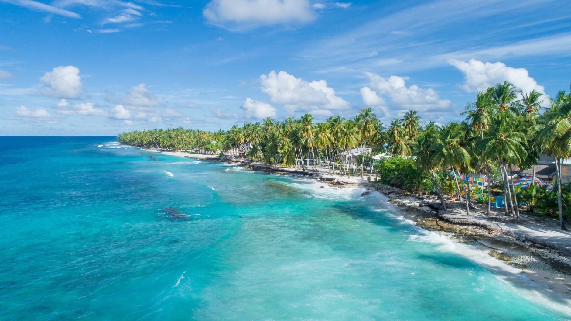 Aerial view of a tropical beach with turquoise waters, white sand, and lush palm trees under a bright blue sky with scattered clouds. The serene coastline features gentle waves and a few buildings nestled among the greenery, perfect for capturing with an Instagram scheduler | plannthat.com