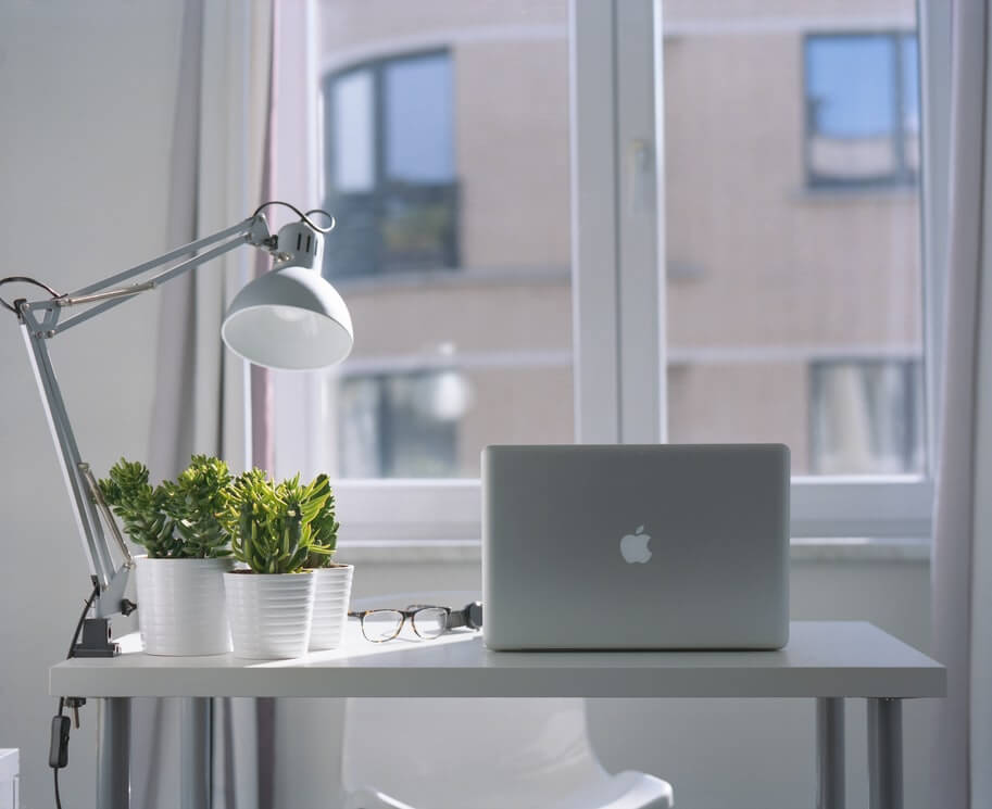 A minimalist desk with a laptop, potted plants, a desk lamp, and glasses, positioned in front of a window with a view of a building, ideal for using a social media scheduler | plannthat.com