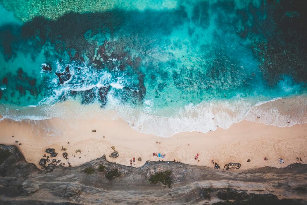 Aerial view of a serene beach with turquoise waters, gentle waves, and a sandy shore. People are relaxing on the beach, surrounded by rocky formations and lush greenery. The vibrant colors highlight the natural beauty of the coastal landscape, perfect for sharing with an Instagram scheduler. | plannthat.com