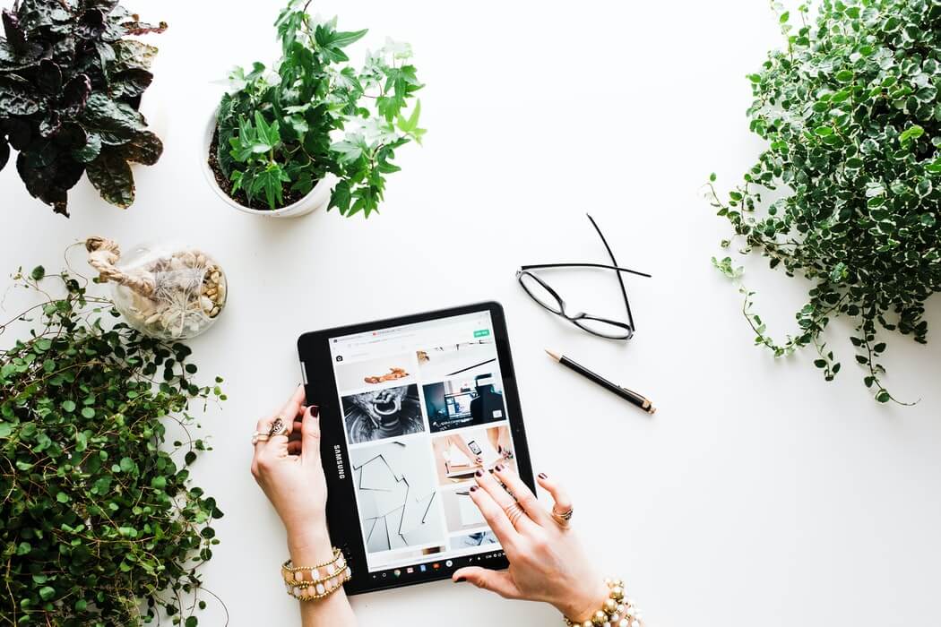 A person browsing on a tablet surrounded by various indoor plants on a white desk, with glasses and a pencil placed nearby, using a social media scheduler | plannthat.com