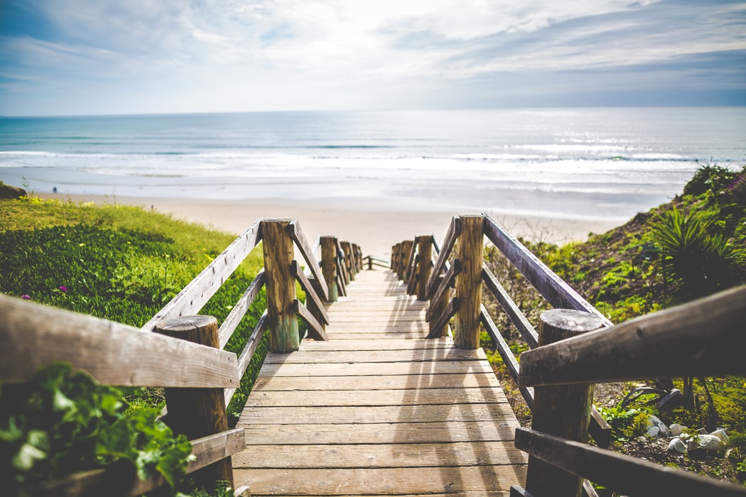 A wooden boardwalk with railings leads down to a serene beach with gentle waves under a partly cloudy sky, flanked by greenery, perfect for a social media scheduler | plannthat.com