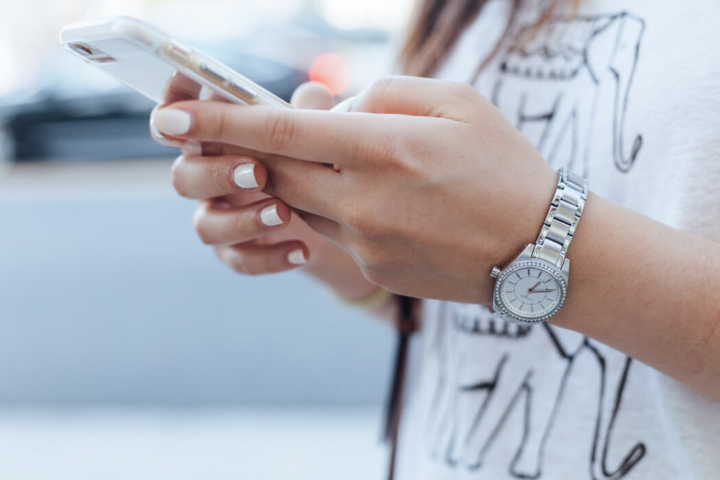 A close-up of a person wearing a silver watch and a white shirt with an elephant design, holding and using a smartphone, emphasizing the hands and phone in a blurred background, ideal for a social media scheduler | plannthat.com
