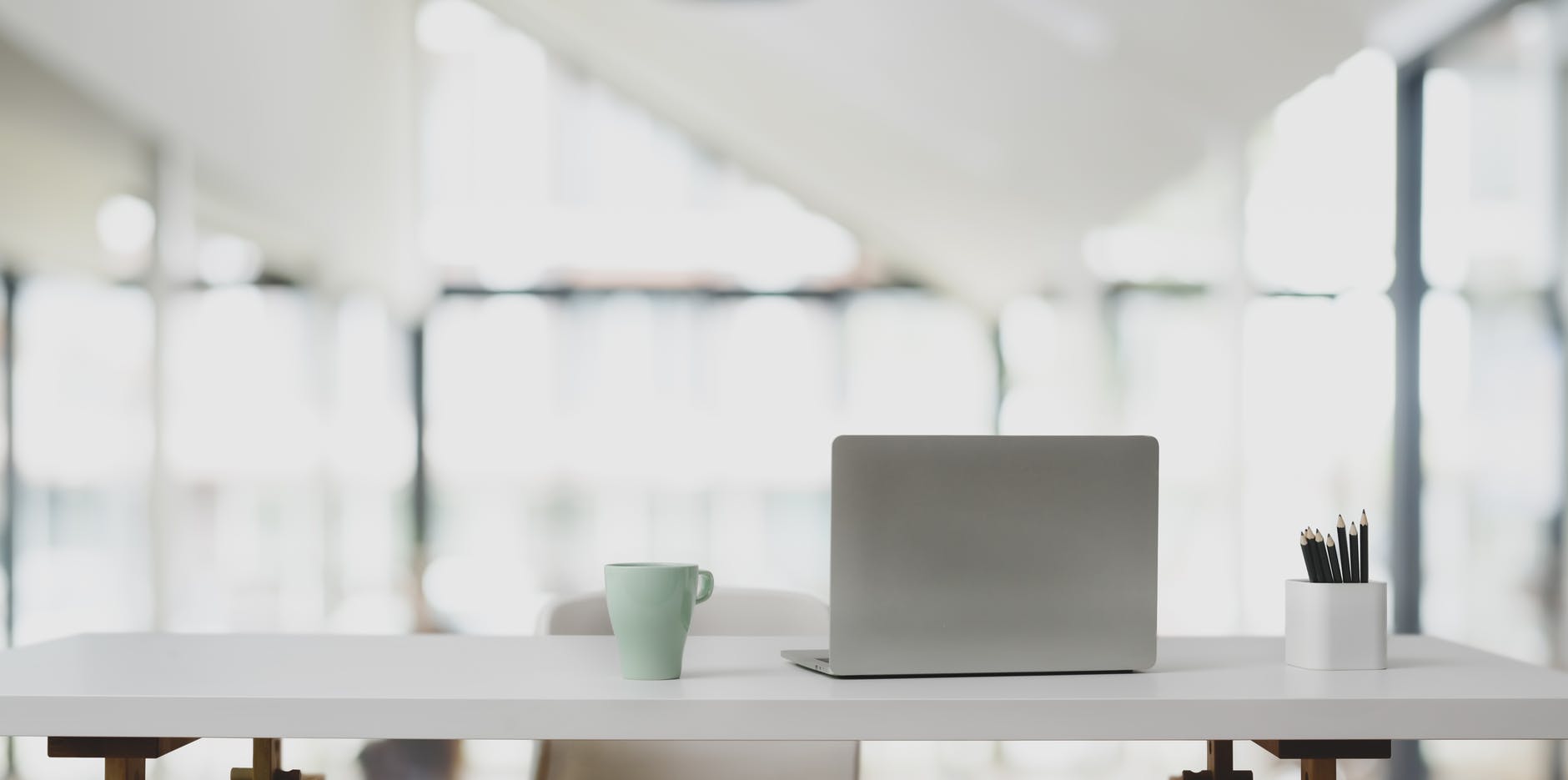 A minimalist workspace featuring a closed laptop, a green coffee mug, and a white pencil holder with pencils on a white desk, with large windows and natural light in the background, creating a bright, airy atmosphere, ideal for using a social media scheduler | plannthat.com